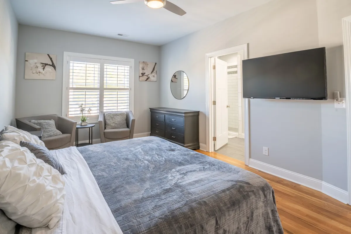 A serene bedroom featuring a plush bed with soft bedding, two comfortable chairs with a small table—perfect for reading or morning coffee—a large wall-mounted TV, a decorative mirror, and tasteful artwork. The room is filled with natural light, creating a calm and private retreat.
