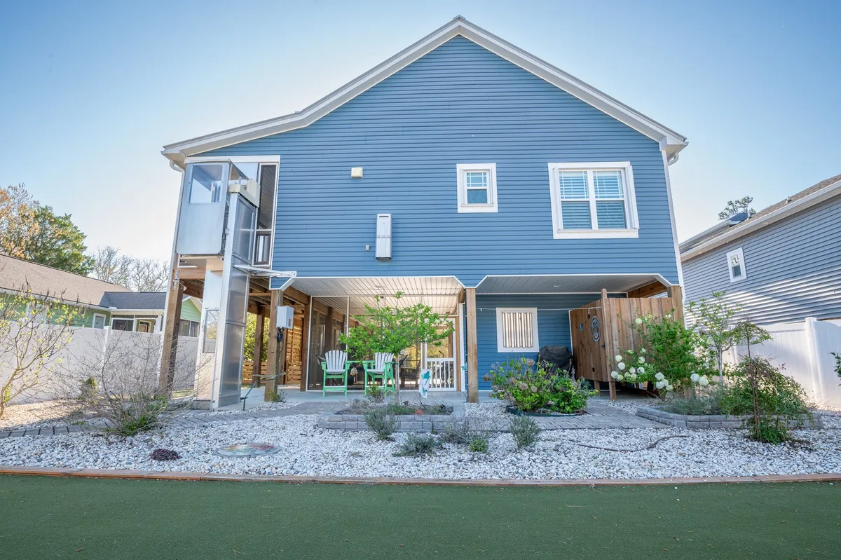 An exterior view featuring the house’s blue siding, lush green plants, and a pair of white chairs on the gravel patio. The casual arrangement suggests a spot for morning coffee, relaxed conversation, or unwinding after a day of exploration.