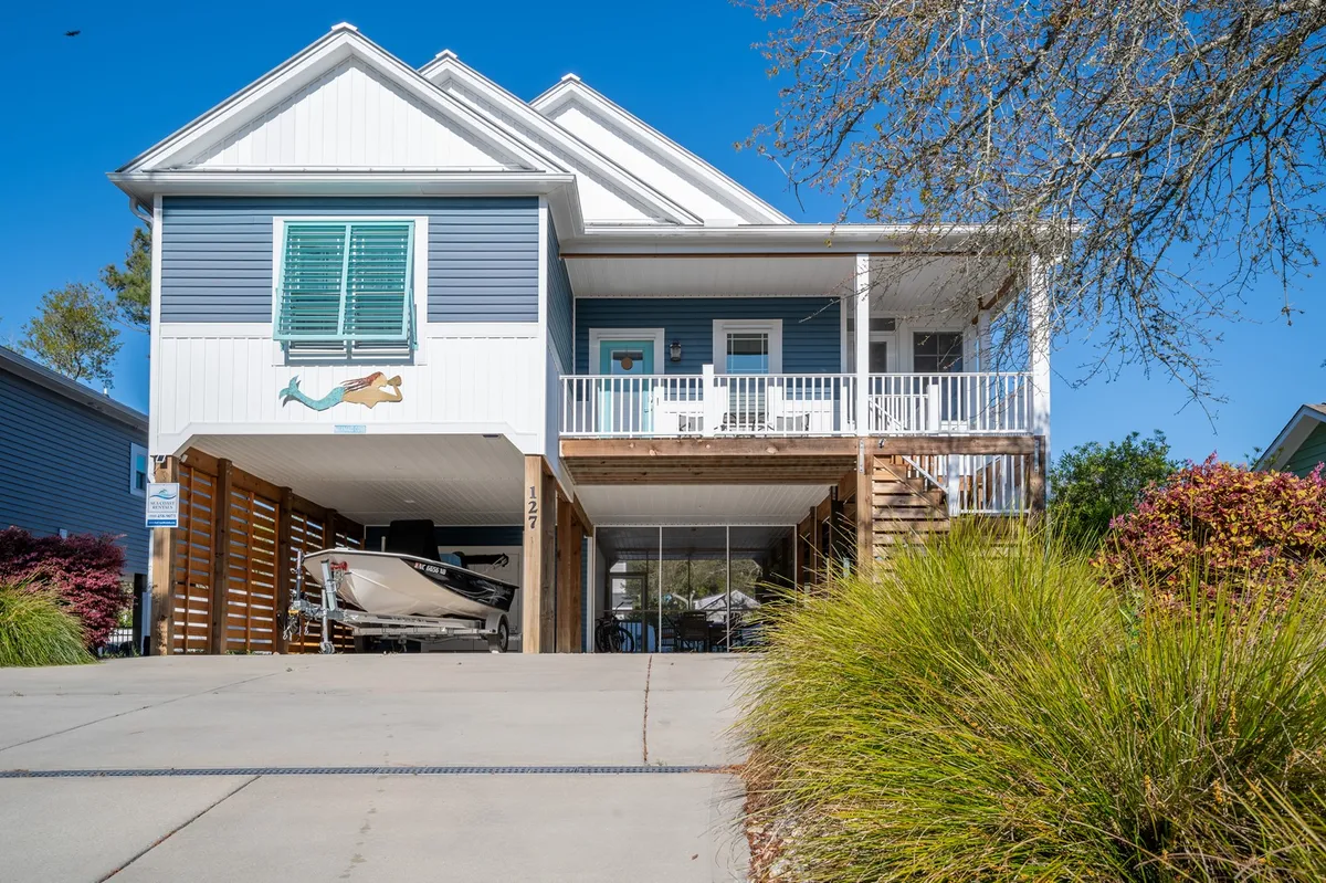 The image shows the front porch of a charming single-family house in Oak Island, North Carolina. The porch is welcoming and ideal for relaxing, while underneath the house, a boat hints at seaside adventures. A colorful mermaid mural adds a playful, coastal touch, and the setting sun creates a warm, inviting glow—perfect for family gatherings and barbecues.