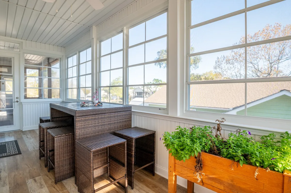 A cheerful sunroom featuring rattan furniture, large windows, and a vibrant planter with flowers, basking in sunlight. The space is perfect for relaxing with a book or enjoying morning coffee while soaking in the views.