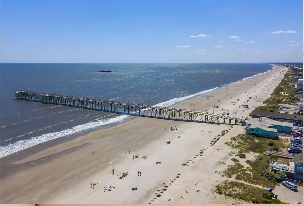 A bustling beach and pier scene with people strolling, sunbathing, and fishing. The shoreline and ocean stretch into the sunset, evoking the sense of relaxation and active, sun-filled days that define coastal living.