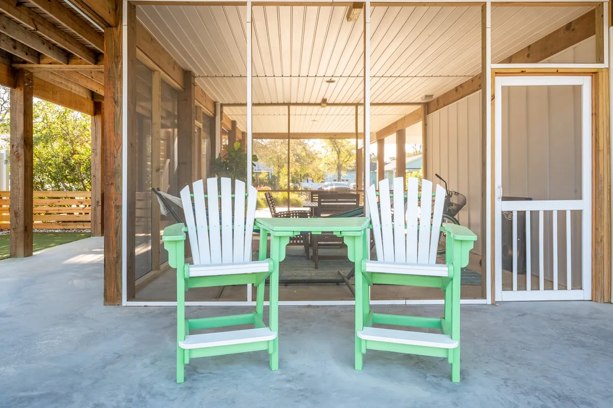 A welcoming screened porch with white and green Adirondack chairs set around a table, with a dining area visible in the background. The space is casual and perfect for relaxing after outdoor adventures, with views of the yard and the promise of shared moments.