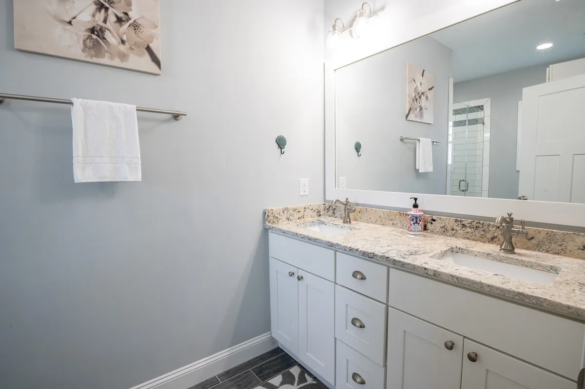 A bright, inviting bathroom featuring a dual sink vanity with a large framed mirror, soft lighting, and decorative wall art. The setup offers ample space for morning routines or winding down after a long day.
