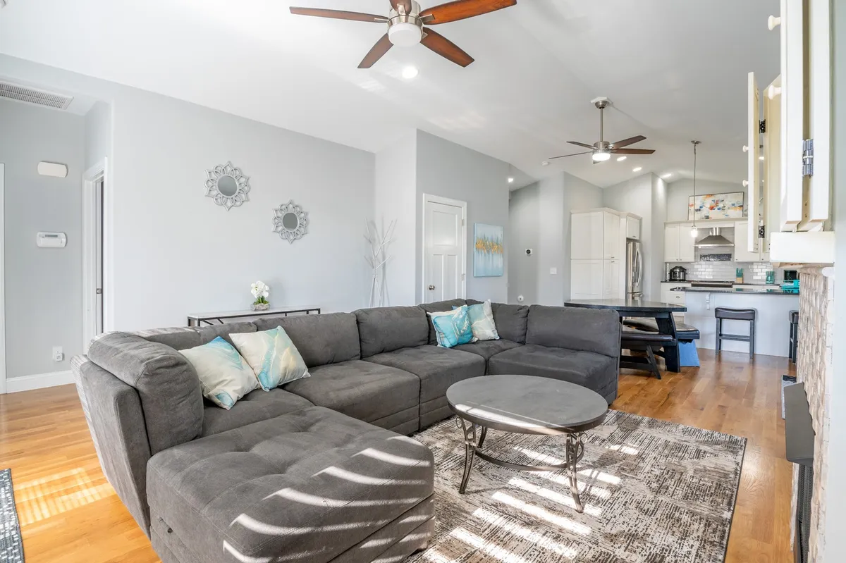 A bright, open-plan living space featuring a plush sectional sofa, a coffee table, decorative mirrors, and a view into the adjoining kitchen area. Natural light fills the room, and the layout encourages mingling and relaxation.