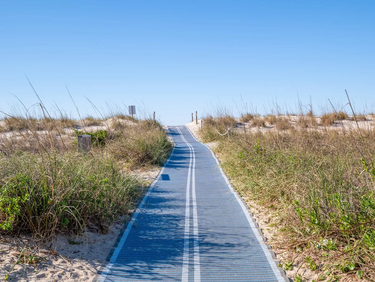 A scenic boardwalk stretches toward the sandy beach, flanked by lush dune grasses and coastal vegetation. The pathway invites viewers to imagine a leisurely walk to the shore, with the natural landscape setting a serene, inviting mood.