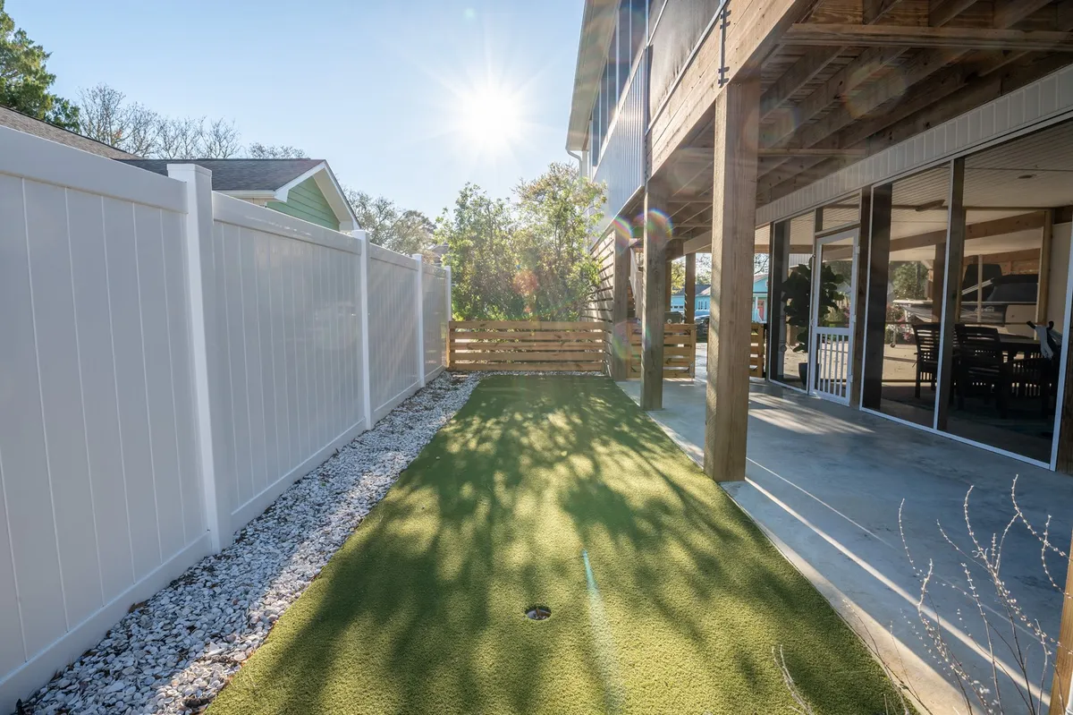 A sunlit outdoor area featuring green turf, a wooden fence, and dappled sunlight streaming through the trees. The space is welcoming and has room for outdoor relaxation, gardening, or small gatherings, evoking a sense of community and enjoyment of nature.