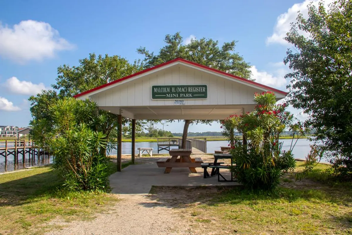 A welcoming community park with wooden picnic tables, a park sign, green surroundings, and a peaceful water view. The setup suggests a space for social gatherings, outdoor play, and connection with nature.