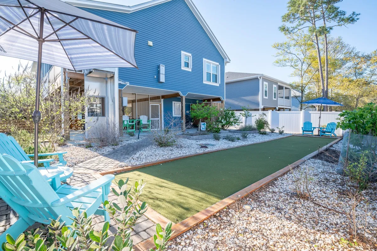 A vibrant backyard scene featuring a lush green lawn, a cluster of blue Adirondack chairs surrounding a patio umbrella, and ample open space for games and gatherings. The area is perfect for outdoor activities such as cornhole, family tournaments, or simply lounging with a cool drink as the sun sets.