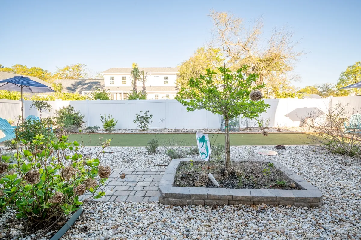 A peaceful garden setting with a green tree, stone pathway, decorative sign, and vibrant flowering plants. The atmosphere is tranquil, ideal for relaxing under umbrellas, watching wildlife, or children playing in a safe, beautiful environment.