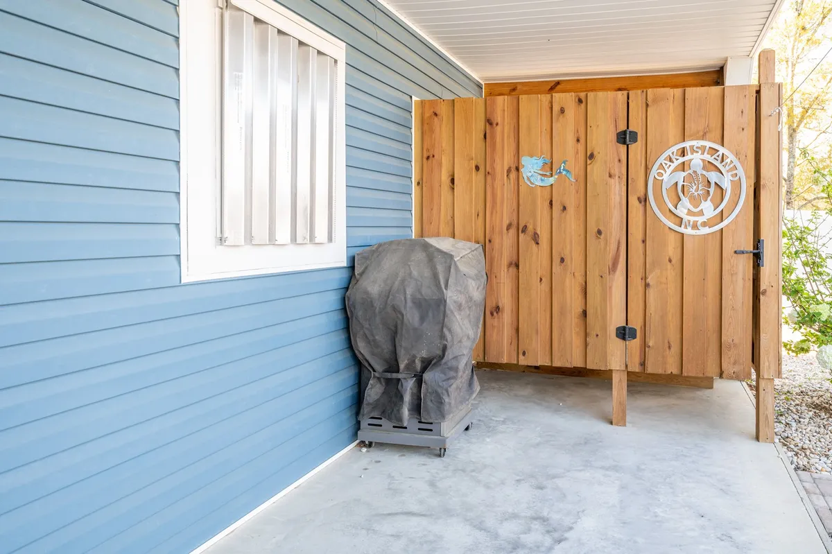 An exterior view featuring blue house siding, a wooden privacy gate, and a covered gas grill. The scene suggests a welcoming outdoor cooking area ideal for sunset barbecues, with space for socializing and enjoying good food in the open air.