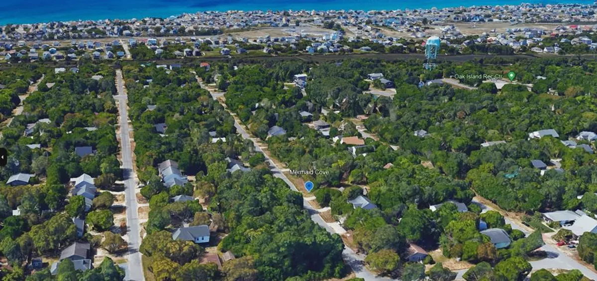 A wide, elevated view of the Oak Island neighborhood, with homes nestled among lush greenery and the expansive ocean visible just beyond. The layout highlights the proximity to the beach and showcases walkable, bike-friendly streets. The scene exudes relaxed coastal living and community connectedness.