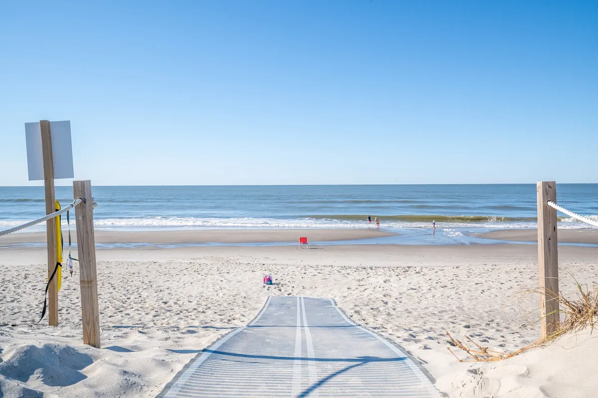 A scenic walkway leading toward the beach, with the ocean and people in the distance enjoying the sun. The scene captures the leisurely, carefree spirit of seaside living and invites viewers to imagine joining in beach activities or local events like fireworks or concerts.