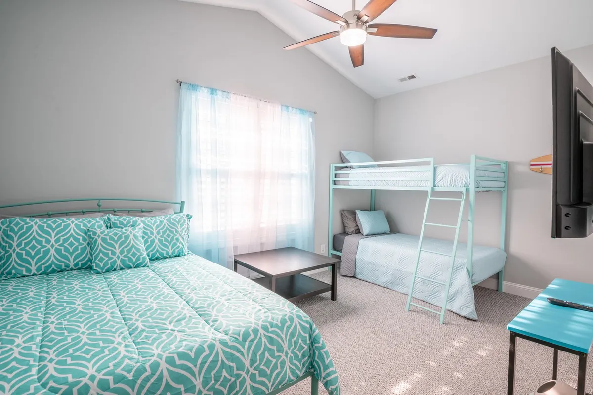A cheerful guest bedroom with a double bed and a twin/twin bunk bed, light blue curtains filtering natural light, a ceiling fan overhead, and a TV. The arrangement shows versatility, ideal for families with children.