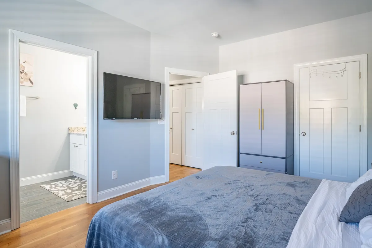 A modern bedroom with a gray-blanketed bed facing a wall-mounted TV, an open closet door, and an entrance leading to a bathroom. The space feels cozy and functional, blending comfort and convenience.