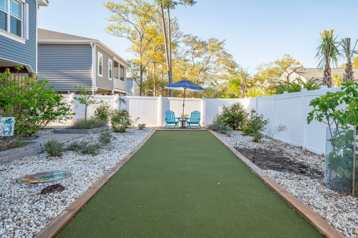 A cheerful backyard scene featuring green artificial turf, blue chairs, a white fence, lush garden plants, and a large umbrella, creating an inviting and secure outdoor space for pets and owners to relax or play together.
