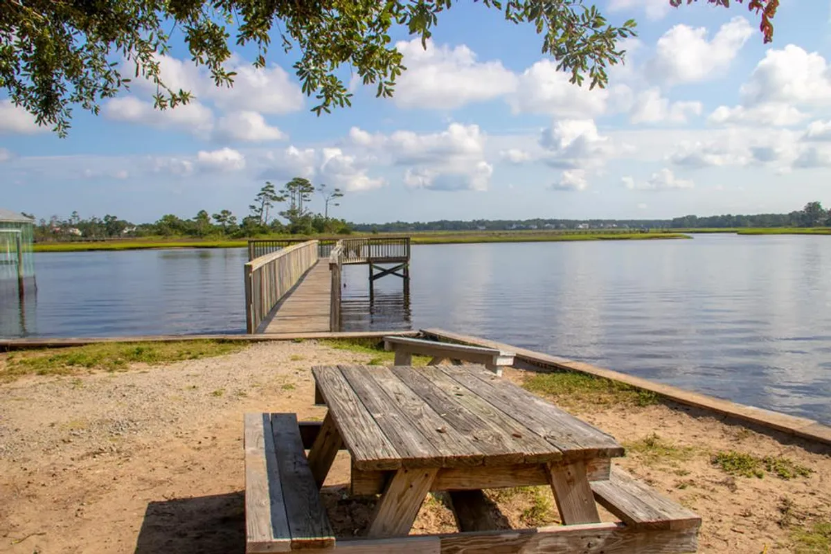 A wooden picnic table sits on a dock overlooking a peaceful waterfront, surrounded by trees. The tranquil setting suggests opportunities for outdoor meals, relaxing, and enjoying coastal wildlife or water-based activities like fishing and kayaking.